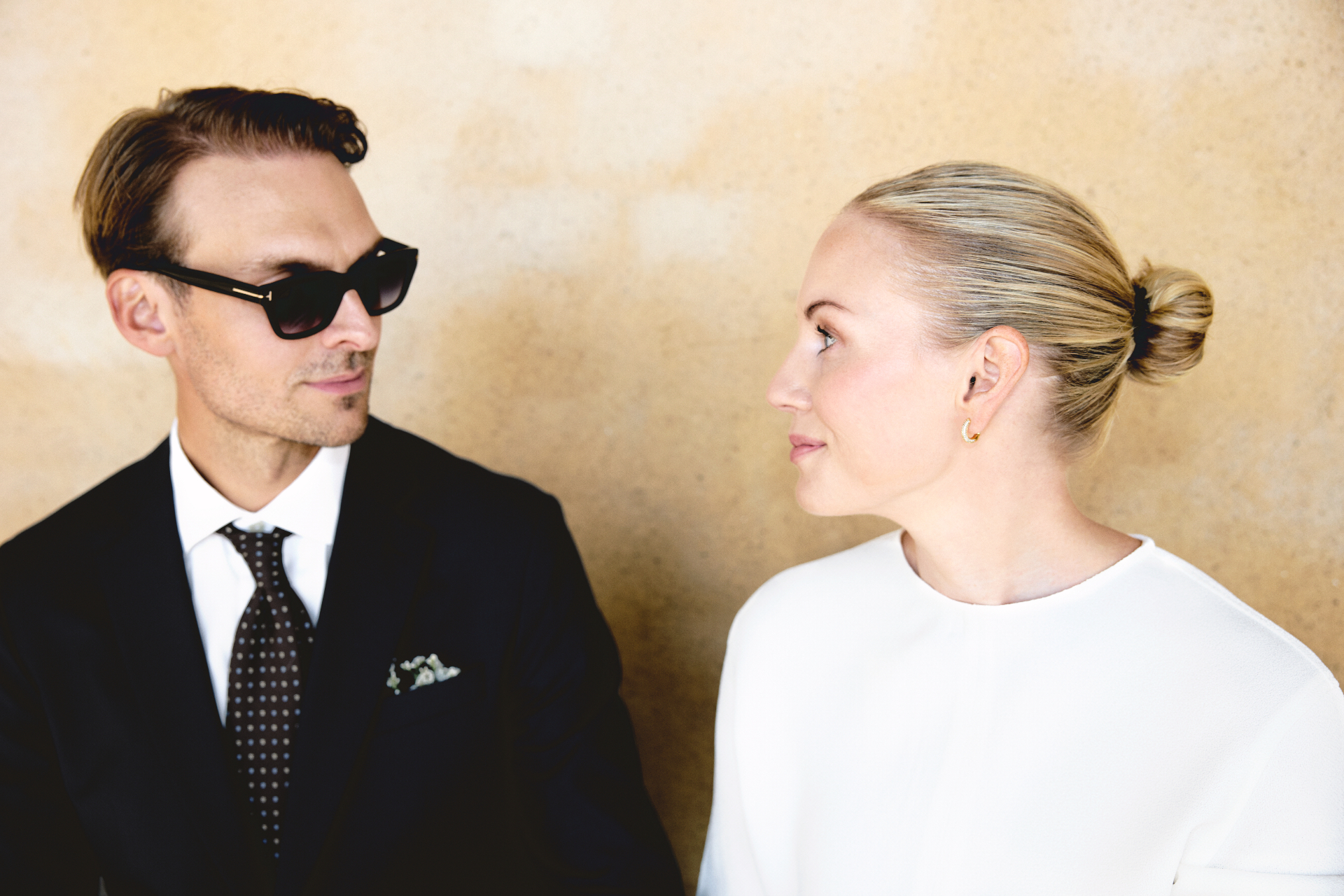 Simon in sunglasses looking at Elin against the ochre wall